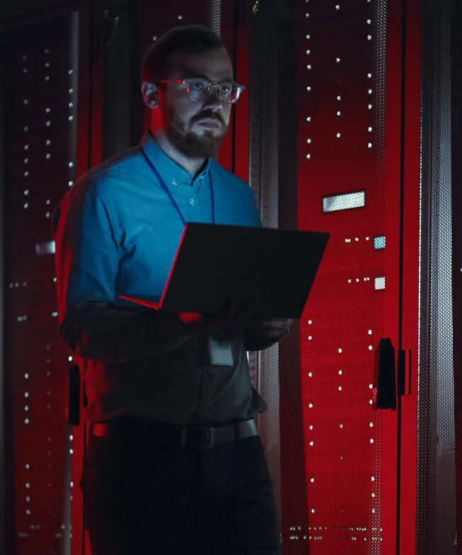 IT professional with a laptop standing beside red-lit server cabinets in a dark data center