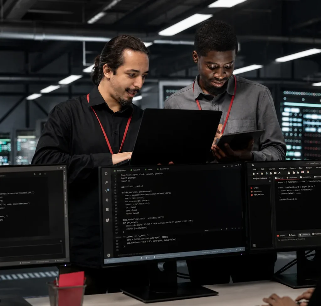 Two IT specialists reviewing code on a laptop and tablet in a network operations center