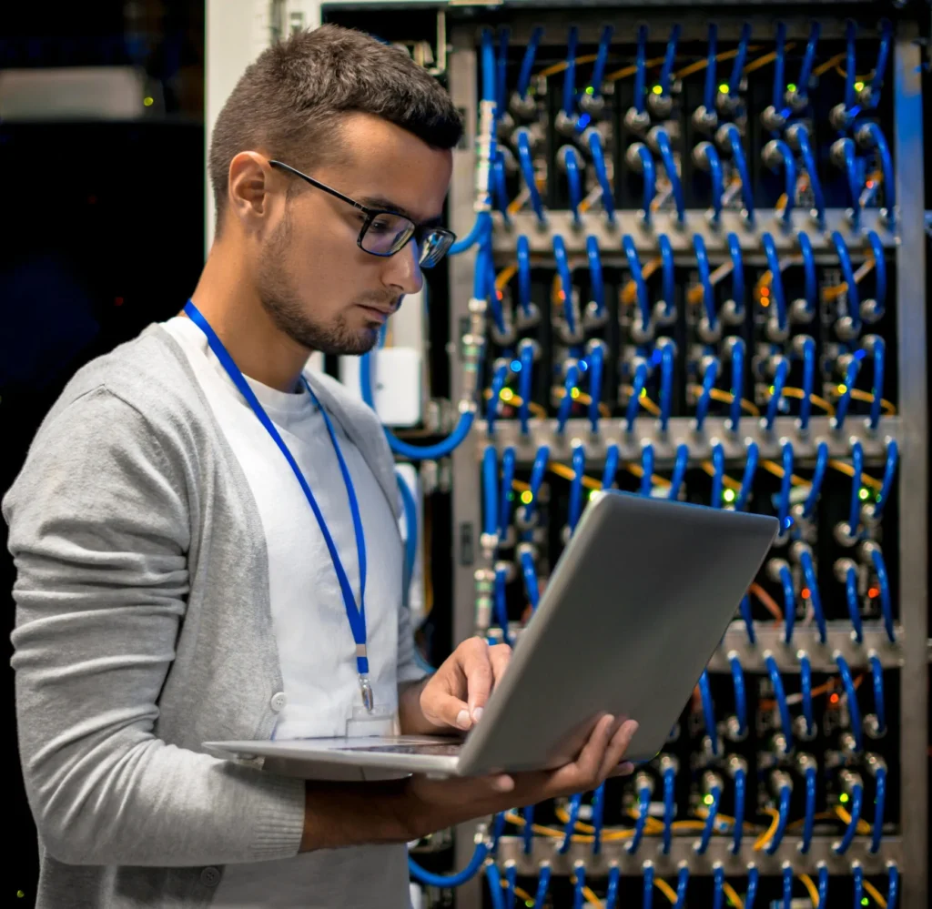 Network engineer with a laptop inspecting a rack of blue patch cables in a server room